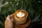 Woman is holding Glass of cappuccino with latte art on saucer by tropical plant in front.