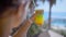 Woman is holding glass with cocktail in background of resting area of a hotel near sea in sunny weather, making small