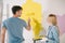 Woman holding roller tray while boyfriend painting wall in yellow with paint roller