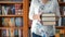 Woman holding a stack of large books with a large bookcase full of books on her back.