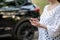 A woman holds a smartphone on the background of a modern car. a business woman stands with a phone by the car