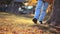 A woman in jeans and boots walks up to a tree, leaning on it and swinging her leg. Legs close up. Autumn Park in the background