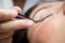 Woman lies on eyelash extension procedure in a beauty salon. Lashmaker holds tweezers with a bunch of artificial