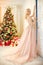 Woman in a long cream-coloured dress, standing near the Christmas tree and the door. Luxurious blonde in evening dress celebrate