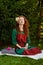 A woman with long curly red hair and closed eyes is sitting serenely on the grass on a blanket and meditating, holding rose petals