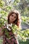 Woman with long hair over branches with white blossom and green leaves looking at camera illuminated with sunshine