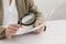 Woman looking at document through magnifier at wooden table, closeup. Searching concept