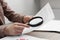 Woman looking at document through magnifier at wooden table indoors, closeup. Searching concept