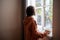 Woman looking at window drinking tea coffee on kitchen at home, rear view.