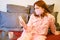 A woman looks at the phone while sitting on a red bed in quarantine. Girl in a makeshift protective mask isolated during