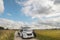 A woman looks up at the sky, a man sits on the step of a car, at a motorhome on a country road, in the middle of fields