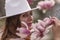 Woman magnolia flowers, surrounded by blossoming trees., hair down, white hat, wearing a light coat. Captured during