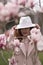 Woman magnolia flowers, surrounded by blossoming trees., hair down, white hat, wearing a light coat. Captured during