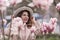 Woman magnolia flowers, surrounded by blossoming trees., hair down, white hat, wearing a light coat. Captured during