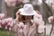 Woman magnolia flowers, surrounded by blossoming trees., hair down, white hat, wearing a light coat. Captured during
