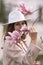 Woman magnolia flowers, surrounded by blossoming trees., hair down, white hat, wearing a light coat. Captured during