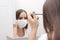 Woman in medical mask does makeup, mascaras eyes at home in the bathroom