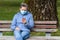 Woman with a medical mask on her face sits on a park bench and looks at the phone, quarantine