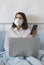 Woman with a medical mask on her face works or studies from home, sits in her bedroom in bed with a laptop and smartphone