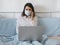 Woman with a medical mask on her face works or studies from home, sits in her bedroom in bed with a laptop and smartphone