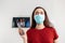 A woman in medical mask holding a remote control from the TV. In the background a white wall with TV monitor. Close up. The