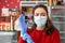 A woman in a medical mask puts on rubber gloves, preparing for a work shift. In the background-shelves and shop windows. The