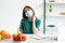 Woman in medical mask sitting at table with fruits, vegetables and pills and scratching neck at home