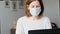 Woman in medical mask working at home office on laptop, confused face, self isolation