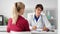 Woman medical worker in glasses and coat talking with girl patient taking note during consultation