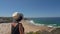 Woman in a middle-aged hat stands on a mountain slope and admires the beautiful view of the Portuguese beach Odeceixe in