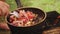 A woman mixes chopped sausage with mushrooms, tomatoes and onions in a frying pan on the coals.