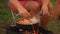 A woman mixes eggs with vegetables and sausage with a spoon in a frying pan standing on the coals.