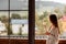 Woman in the morning holding a cup of tea or coffee and looking at the mountains standing near the window