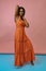 woman in orange dress posing with hands behind head and on neck on blue and pink studio background