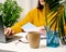 A woman in an orange sweater sits at a white table and works at a laptop. Freelancer at remote work, salesperson`s home office