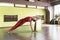 A woman performs the exercise kamatkarasana, dancing dog pose, stands on a mat in the studio, trains in a red sports jumpsuit