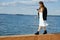 Woman with phone in hand walks across pier in white dress and jacket stares at screen