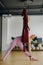 A woman in pink clothes does yoga on a suspended burgundy hammock in a bright gym