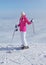 Woman in pink jacket, ski poles and skis, standing at the edge of hill, only clouds below, looking back, smiling