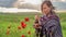 Woman in poncho with singing bowl sits on flower field under cloudy sunset sky.