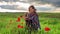 Woman in poncho with singing bowl sits on flower field under cloudy sunset sky.