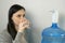 Woman is pouring water into glass from an automatic water cooler and drinking.