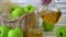 A woman pours apple juice from a jug into a glass. Nearby are apples on the table