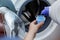 Woman pours liquid washing gel into plastic cap against background of drum of steel-colored washing machine. A girl