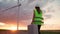 Woman Professional Ecology Engineer in uniform and helmet with special equipment in hand goes service windmill on beautiful sunset