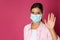 Woman in protective mask showing hello gesture on pink background, space for text. Keeping social distance during coronavirus