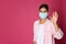 Woman in protective mask showing hello gesture on pink background, space for text. Keeping social distance during coronavirus