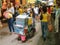 Woman pushes bottled drinks on cart through lane between market stalls and people