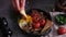 woman puts tasty dish breakfast onto dark background - fried bacon, eggs, mushrooms and tomatoes in ceramic pot