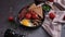 woman puts tasty dish breakfast onto dark background - fried bacon, eggs, mushrooms and tomatoes in ceramic pot